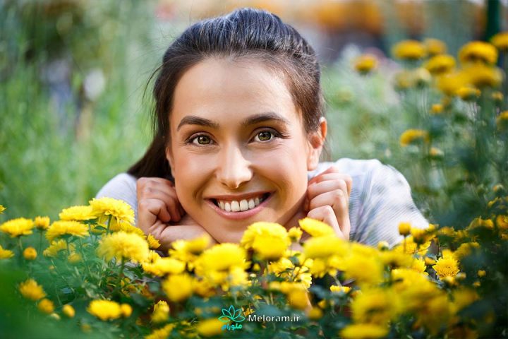 زنی با موهای مشکی که با لبخند و دست بر چانه از میان گل های زرد به روبرو نگاه می کند.